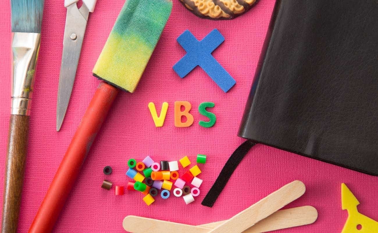 VBS craft supplies, colorful foam letters, pom-pom sticks, black notebook, paintbrush, scissors, and wooden sticks on pink background for Vacation Bible School activities.