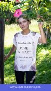 Blessed woman in a white floral T-shirt with biblical verse about purity and seeing God, standing outdoors among greenery, holding a branch with pink flowers, promoting Christian faith and biblical inspiration.