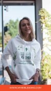 Comfortable woman wearing a white Christian hoodie with religious quote, standing outdoors near a church or chapel, promoting Christian music and faith-based prayer through My Bible Song.