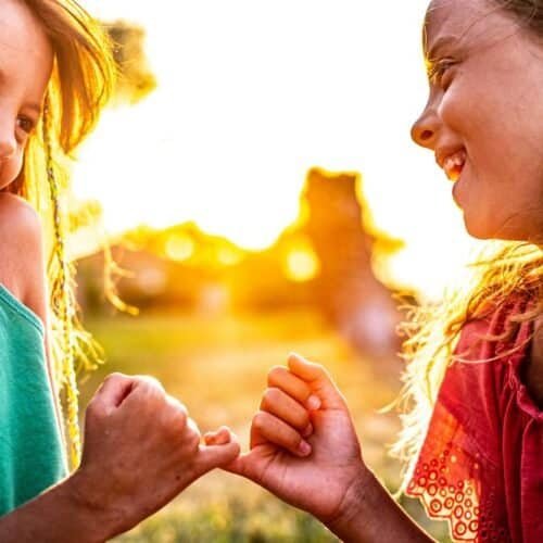 Joyful girls arm wrestling outdoors during sunset, emphasizing faith-based friendship and happiness, inspiring children’s spiritual growth and love for God through song.