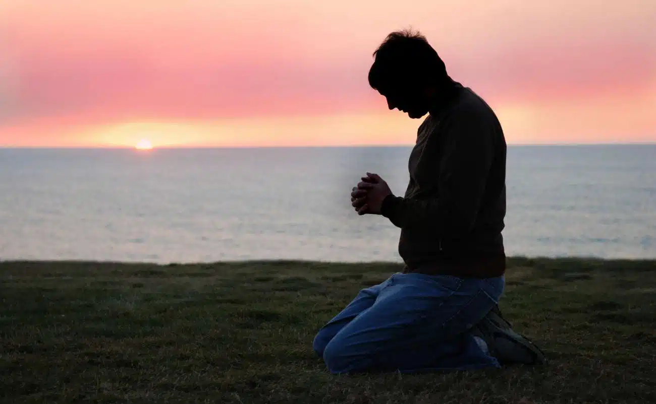 Praying man kneeling by the ocean at sunset, reflecting faith and devotion, with the serene sea and colorful sky in the background, emphasizing spiritual connection and prayer.