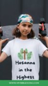 Holding skateboard at shoulder, girl wearing "Hosanna in the Highest" t-shirt, Christianity faith symbol, modern religious youth, outdoor street style.