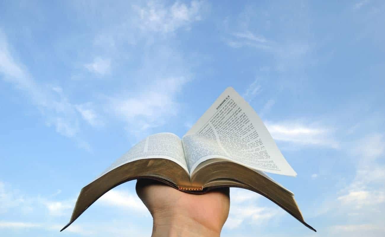 Bible open in hand against blue sky with clouds, symbolizing faith and spiritual growth, promoting biblical teachings and Christian devotion.