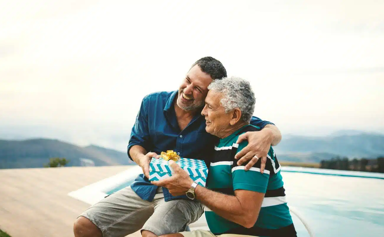 Generates a photo of two men celebrating and holding a gift, outdoors, with a scenic mountain background, capturing joy, gratitude, and connection, suitable for promoting faith, family, and gift-giving themes related to Bible songs.