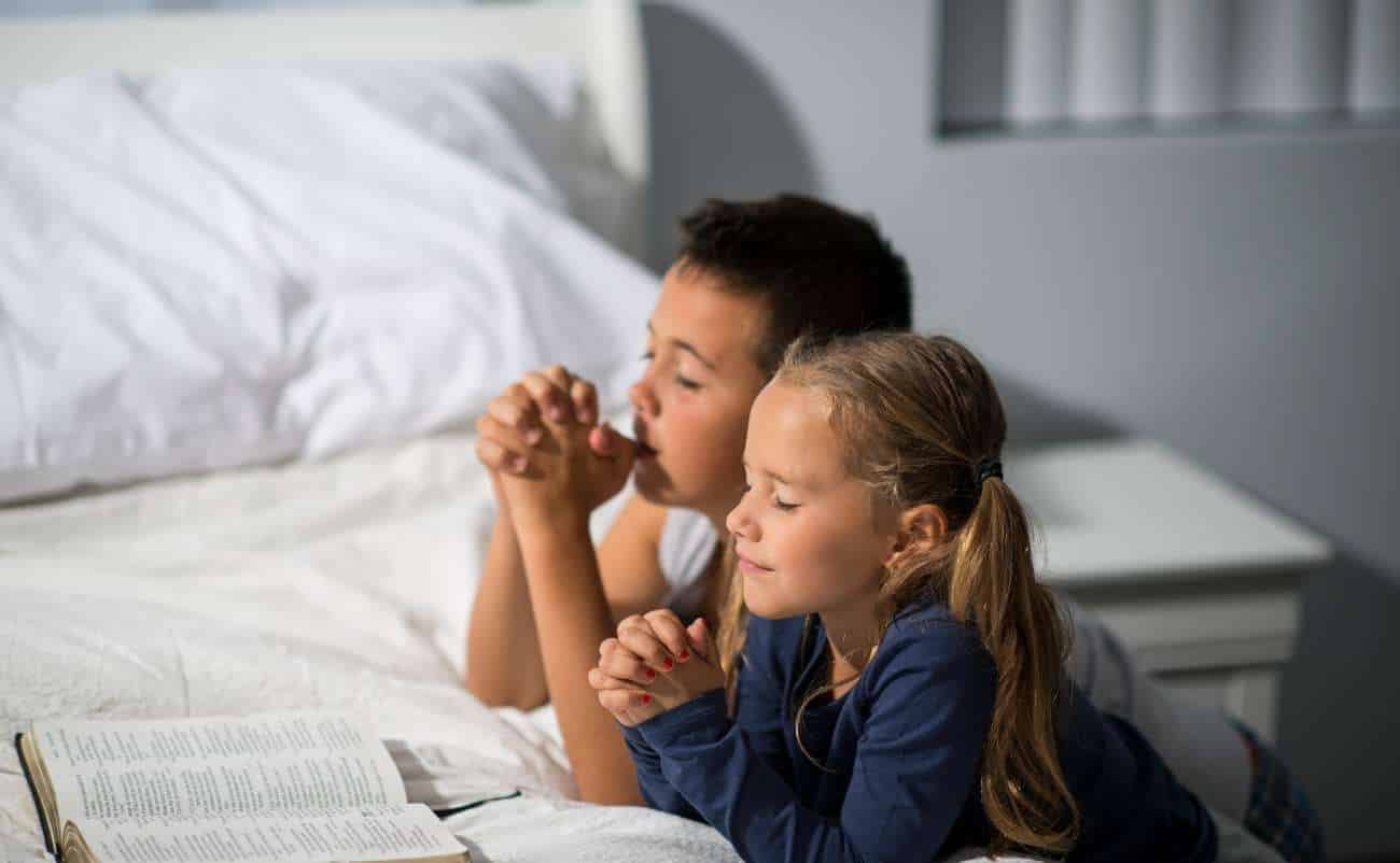 Children praying with closed eyes and folded hands, reading the Bible in a peaceful bedroom setting.
