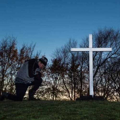 Start Your Day with Faith: Powerful Christian Morning Prayers for a Blessed Morning My Bible Song 2026 Kneeling person praying by a white cross on a grassy hill, with leafless trees and a clear evening sky, symbolizing faith, spirituality, and Christian devotion, suitable for Bible song and worship themes.