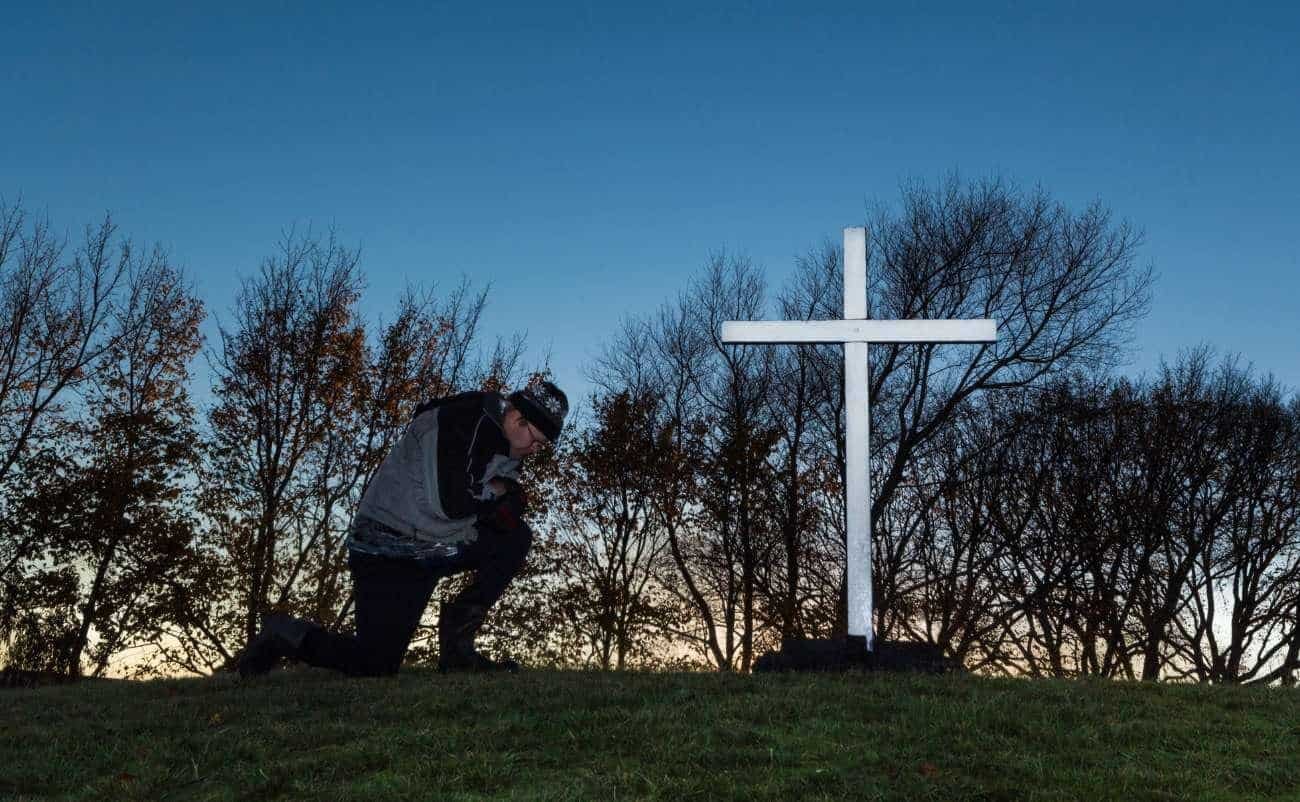 Kneeling person praying by a white cross on a grassy hill, with leafless trees and a clear evening sky, symbolizing faith, spirituality, and Christian devotion, suitable for Bible song and worship themes.