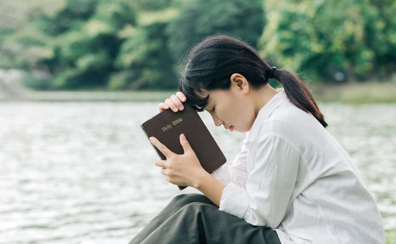 A young woman praying outdoors while holding a Holy Bible close to her forehead, depicting faith, spirituality, and devotion. She is seated by a peaceful river, surrounded by lush greenery, reflecting tranquility and spiritual connection.