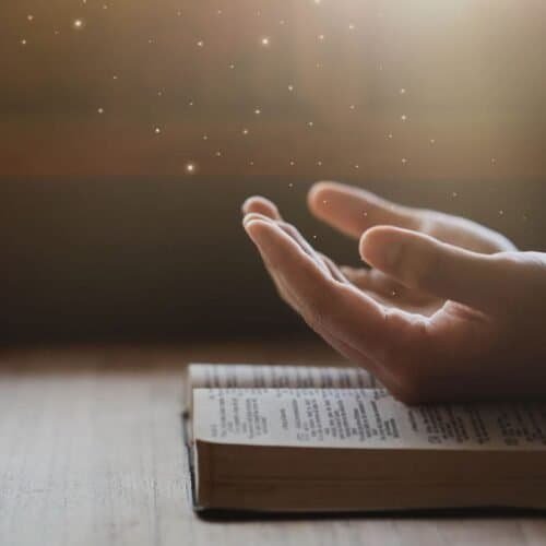 Inspiring hands hovering above an open Bible with glowing light and star-like particles, representing faith and spirituality through biblical songs and Christian worship.