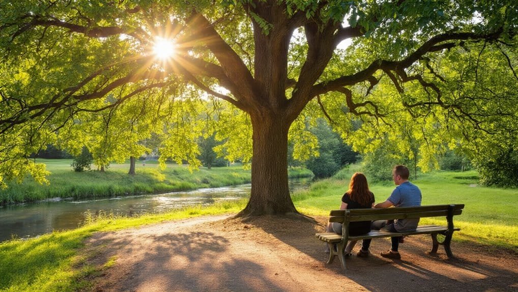 A peaceful outdoor scene with a couple sitting on a park bench under a large, leafy tree by a calm river during sunset, perfect for inspiration and reflection.