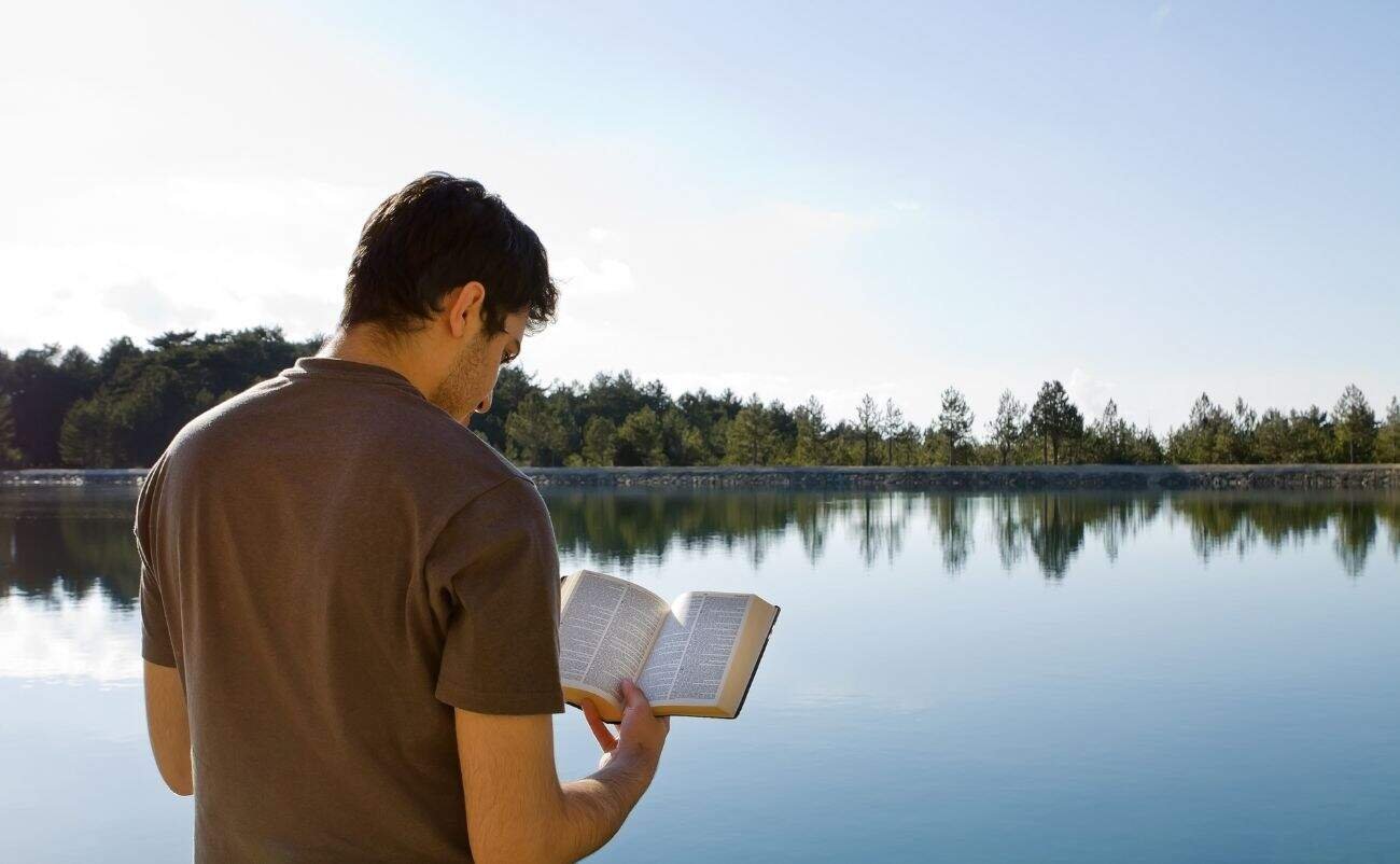 Quiet man reading Bible next to tranquil lake in nature, practicing faith, prayer, spirituality, biblical study, outdoor worship, spiritual reflection, peaceful outdoor scene.