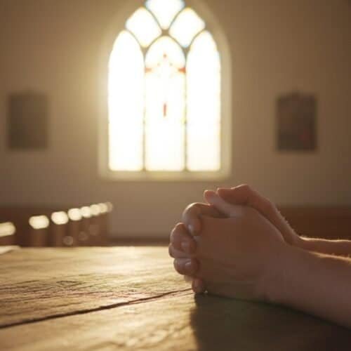 Praying hands at church altar with sunlight streaming through stained glass windows, symbolizing faith, worship, and devotion in Christian practice.